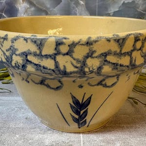 May include: A cream-colored ceramic mixing bowl featuring a blue sponge-painted rim and a hand-painted blue wheat stalk motif on the front. The bowl has a wide, circular opening and a smooth, glazed finish throughout its exterior and interior surface.