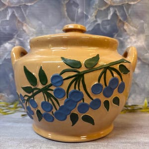May include: A tan ceramic sugar bowl with a matching lid and two side handles. The exterior features a raised, hand-painted design of blue berries on green leafy branches. The glaze has a smooth, glossy finish with a speckled texture throughout.