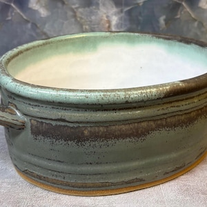 May include: A ceramic bowl with two side handles, featuring a smooth white interior and a textured exterior glazed in shades of sage green and dark brown. The base of the bowl is unglazed, revealing a natural tan clay color along the bottom edge.
