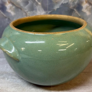May include: A round, sage green ceramic bowl with a smooth, speckled glaze finish. The bowl features a wide, open top with a thick, unglazed tan rim and two small, integrated side handles extending from the upper body of the vessel.