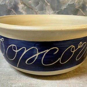 May include: A large, round ceramic bowl with a wide, flared rim. The upper portion is a speckled cream color, while the lower section features a deep blue band with the word Popcorn written in a white, cursive, hand-painted script style.