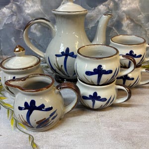 May include: A ceramic tea set featuring a teapot with a lid, a lidded sugar bowl, a creamer, and three cups. Each piece is off-white with hand-painted blue floral motifs and brown rim accents. The teapot has a curved handle and spout.