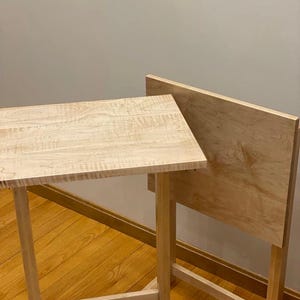 May include: Two light-colored wooden snack tables with rectangular tops and simple four-legged frames. The wood features a visible natural grain pattern. One table is positioned horizontally, while the other is standing vertically behind it.