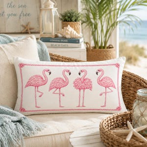 May include: A rectangular lumbar cushion featuring a white fabric cover with a pink rope-style border and a graphic print of four pink flamingos in various standing poses. The design is centred on the front of the cushion.
