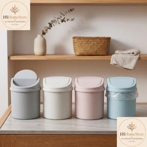 Puede incluir: Cuatro cubos de basura cilíndricos pequeños con tapas basculantes, dispuestos en fila. Los cubos son de color gris claro, blanco roto, rosa suave y azul claro. El cubo azul claro tiene una bolsa de plástico transparente en el borde.