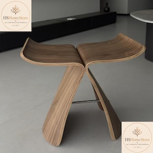May include: A butterfly stool crafted from bent plywood with a visible wood grain finish. The seat consists of two symmetrical, curved panels that meet in the center, supported by a thin metal rod connecting the two legs at the base.