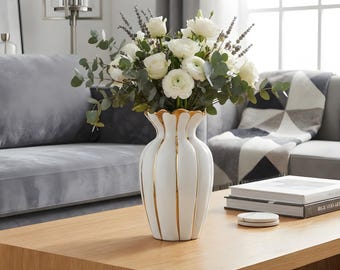 May include: A white ceramic vase with vertical gold-painted grooves containing a bouquet of white roses, ranunculus, lavender sprigs, and eucalyptus leaves. The vase has a scalloped rim and a rounded body, standing on a wooden surface.
