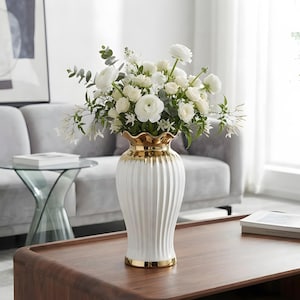 May include: A white ceramic vase with vertical fluted ridges and a metallic gold rim at the top and base. The vase is filled with a bouquet of white ranunculus flowers, small white blossoms, and green eucalyptus leaves arranged in a rounded shape.