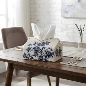 Könnte beinhalten: Ein rechteckiger Taschentuchbox-Behälter aus Keramik mit cremefarbener Basis und einem detaillierten blauen Blumenmuster. Das Design zeigt verschiedene blühende Blumen und Blätter auf der Vorder- und Seitenfläche, aus der oben weiße Taschentücher ragen.