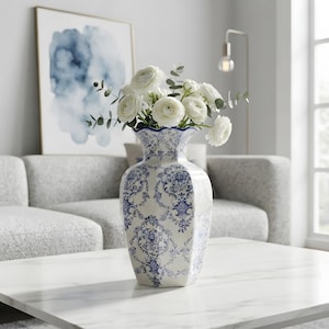 Peut inclure: Un vase en céramique blanche orné d'un motif floral damassé bleu avec un bord festonné, contenant un bouquet de renoncules blanches et de tiges d'eucalyptus vertes. Le vase a une forme hexagonale et repose sur une surface en marbre blanc.