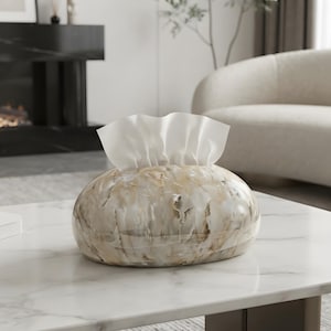 May include: An oval-shaped tissue box cover featuring a glossy, marbled finish with swirling patterns of cream, beige, and white. A stack of white tissues is pulled through the central opening at the top of the smooth, rounded container.
