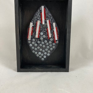 May include: A framed abstract art piece. The artwork features a dark gray, textured, diamond-shaped design with red and silver accents. The bottom portion of the diamond is covered in small, gray, spherical beads. The frame is black.