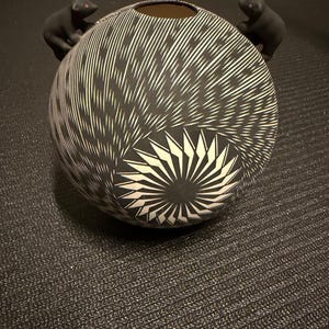 May include: A spherical ceramic vessel featuring a matte black finish with intricate white etched line patterns radiating from a central starburst design. Two small, sculpted black bear figures with subtle pink accents are attached to the rim.
