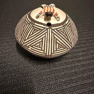 May include: A round ceramic vessel with a cream-colored base featuring black geometric triangular patterns. The top of the vessel has a small circular opening and a sculpted figure with a black head and orange and white striped body.