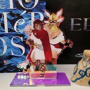 May include: A double-sided acrylic standee featuring two illustrated characters in ancient-style attire. One character wears a white tunic, red cloak, and holds a sword. The other wears a gold winged helmet and white robes. The standee has a purple base.