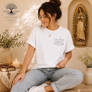 May include: A white short-sleeved t-shirt featuring black cursive text on the left chest that reads: Hecha con propósito, tejida por Dios. Amada por él, y creada para brillar. Isaías 43:13. The shirt is paired with light blue denim jeans.