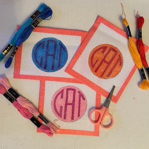 May include: Three square needlepoint canvases with coral borders, each featuring a circular monogram with the letters CAT. Included are six skeins of embroidery thread in blue, yellow, red, and pink, along with a pair of small pink scissors.