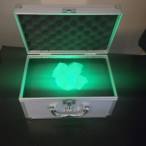 Op de afbeelding: Een zilverkleurige metalen koffer met een scharnierend deksel en een handvat in het midden. De binnenkant is voorzien van donkere schuimvulling en een heldere groene lichtbron die een doorschijnend, onregelmatig gevormd object verlicht.
