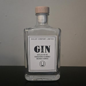 May include: A clear glass bottle with a light wood stopper. The white label features black text reading Shelby Company Limited, GIN, Distilled for the eradication of seemingly incurable sadness, and a horseshoe icon centered at the bottom.
