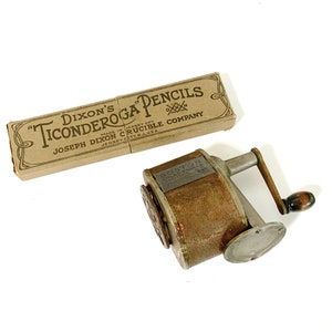 Puede incluir: Un sacapuntas antiguo con un mango de madera y una caja de lápices "Ticonderoga". El sacapuntas es de metal con un aspecto desgastado y envejecido. La caja de lápices es de color beige con texto negro.