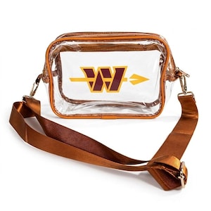 May include: A clear rectangular crossbody bag with brown trim and a matching brown adjustable strap. The front of the bag features a burgundy and gold logo consisting of a stylised letter W intersected by a horizontal arrow pointing to the right.