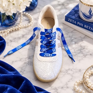 May include: A white sneaker featuring a lace overlay with subtle sequins and bright blue satin shoelaces printed with the Greek letters ZΦB. The shoe has a light-colored sole and a padded interior lining, showcasing a clean and detailed design.