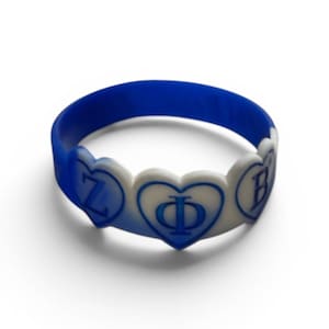 Puede incluir: Una pulsera de silicona azul con una serie de detalles en forma de corazón blanco. Cada corazón contiene una letra griega azul, específicamente Zeta, Phi y Beta, mostrada en una fuente clara y negrita. La pulsera es flexible y circular.