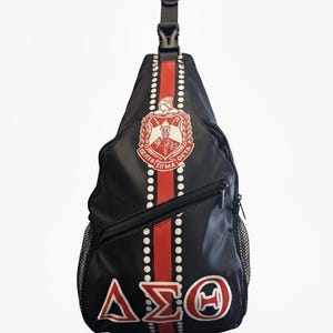Może przedstawiać: Czarny plecak na jedno ramię z centralnym pionowym czerwonym paskiem w białe kropki. Torba posiada czerwono-biały herb u góry oraz greckie litery Delta Sigma Theta w kolorze czerwonym i białym u dołu. Zawiera czarny pasek z klamrą.