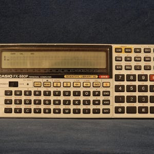 May include: A Casio FX-880P personal computer with a rectangular LCD screen displaying the number 1. The device features a QWERTY keyboard layout, a numeric keypad on the right, and various function keys. The casing is light grey with black keys.