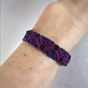 May include: A handmade macrame bracelet in shades of purple and red. The bracelet features an intricate woven design with a repeating pattern. The bracelet is worn on a wrist, showcasing the detailed craftsmanship and color combination.