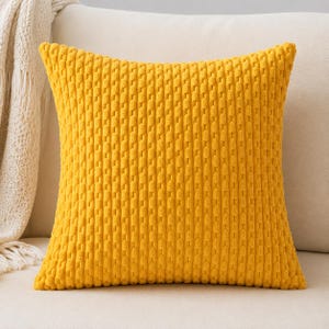 Puede incluir: Un cojín cuadrado de color amarillo mostaza vibrante. El cojín presenta una tela texturizada en relieve estilo pana con un patrón repetitivo de costillas verticales en toda la superficie frontal, creando una apariencia suave y táctil.