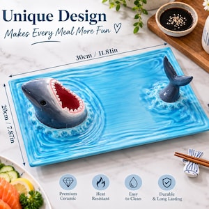 Puede incluir: Una bandeja de servir rectangular de cerámica azul con una cabeza de tiburón tridimensional con la boca abierta en un lado y una cola de tiburón en el otro. La bandeja mide 30 cm por 20 cm y presenta una textura de superficie de agua ondulada.