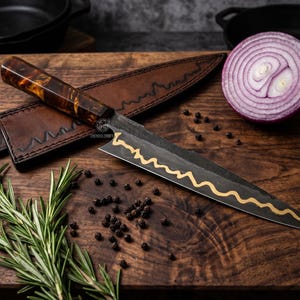 May include: A kitchen knife with a dark, textured blade featuring a gold-colored wavy line, paired with a brown resin handle and a matching leather sheath. The knife is displayed alongside a sliced red onion, rosemary, and black peppercorns.