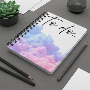 May include: A spiral-bound notebook with a white cover featuring a graphic of soft, pastel pink and blue clouds. The text "To do.." is printed in black cursive script on the upper right side of the cover. The notebook has black wire binding.