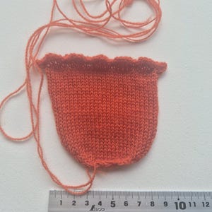 May include: A small, orange knitted pouch with a scalloped top edge and loose yarn strings attached. The pouch is positioned above a metal ruler, showing a width of approximately 3.5 inches across the top opening of the knitted item.