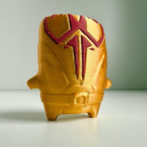 Peut inclure: Une figurine imprimée en 3D avec une finition dorée métallique et un emblème géométrique rouge foncé sur la partie supérieure. L'objet présente un corps cylindrique, de petits bras latéraux saillants et deux jambes courtes à la base, avec des lignes de couche horizontales visibles dues au processus d'impression.