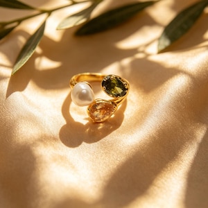 May include: A gold ring featuring an open band design with three distinct stones: a round white pearl, a faceted oval olive green gemstone, and a faceted pear-shaped light amber gemstone, all set in a polished gold-tone metal setting.