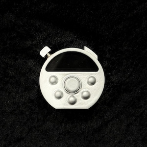 May include: A circular silver-colored handheld device with a matte finish. It features a horizontal black display screen at the top, a large central circular button, and five smaller circular buttons arranged in a semi-circle below the screen.