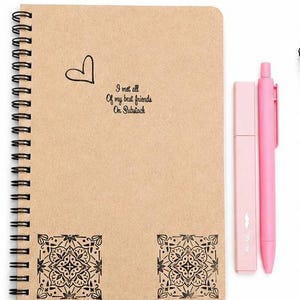 May include: A spiral-bound notebook with a tan cover featuring a black heart drawing, the text "I met all of my best friends on Substack," and two black decorative square patterns at the bottom, accompanied by a pink highlighter and a pink pen.