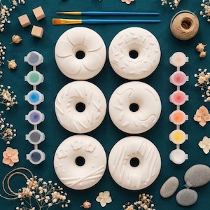 Könnte beinhalten: Sechs weiße, donutförmige Keramikornamente mit verschiedenen Prägemustern, zwei blaue Pinsel, zwei Streifen mit je sechs Farbtöpfen in verschiedenen Farben, drei Holzwürfel, ein Knäuel Garn und getrocknete Blumen auf einem dunklen Untergrund.