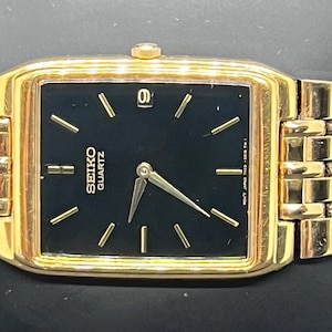 May include: A gold-tone Seiko Quartz wristwatch featuring a rectangular case with a black dial, gold-tone hands, and gold-tone hour markers. The watch includes a date window at the twelve o'clock position and a matching gold-tone metal link bracelet.