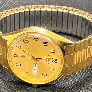 May include: A gold-tone Citizen Quartz wristwatch with a round gold face, black Arabic numerals, and a day-date window at the three o'clock position. The watch features a flexible, expandable metal link bracelet with gold-tone outer links.