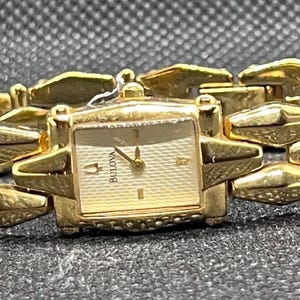 May include: A gold-tone Bulova wristwatch featuring a rectangular face with a textured cream dial, gold hands, and a logo. The watch has a distinctive link bracelet composed of repeating, faceted, diamond-shaped segments with a polished finish.