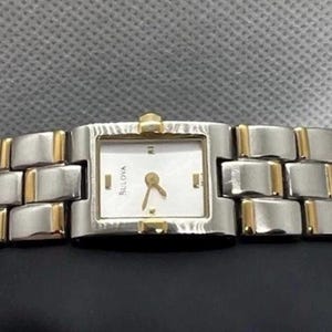 May include: A Bulova wristwatch featuring a rectangular white face with gold-tone hands and markers. The watch has a two-tone link bracelet constructed from silver-tone and gold-tone metal segments, creating a polished and structured appearance.