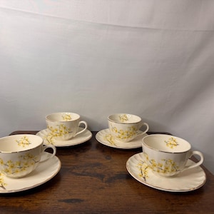 May include: A set of four off-white porcelain teacups and matching saucers, each featuring a delicate yellow floral pattern of blooming branches. The cups have a rounded shape with a gold-trimmed rim, and the saucers have a scalloped edge.
