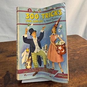 May include: A vintage softcover book titled 300 Tricks You Can Do by Howard Thurston. The cover features a colorful illustration of a magician in a top hat and a person in a wizard hat, surrounded by floating playing cards and a rabbit.