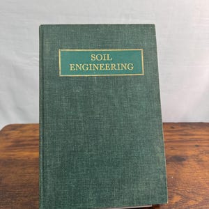 May include: A hardcover book titled Soil Engineering with a dark green textured fabric cover. The front cover features a rectangular inset label with a dark green background and gold lettering that reads Soil Engineering in capital letters.