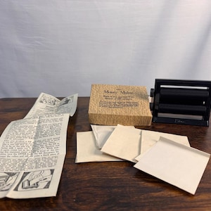 May include: A black plastic money maker magic trick device with rollers, accompanied by a tan cardboard box labeled Money Maker, a printed instruction sheet with illustrations, and several small, blank, rectangular pieces of white paper.