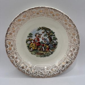May include: A round ceramic plate featuring a central illustration of two figures in period clothing sitting in a landscape with trees. The rim is decorated with an intricate gold and brown floral pattern on a cream background.