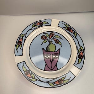 May include: A round ceramic plate featuring a central illustration of a purple cat with a green vase on its head containing red flowers. The rim is decorated with a light blue border, black outlines, and a repeating pattern of red roses and green leaves.